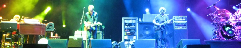 Image of the band Phish with someone playing a brown piano stage left, two guitarists in stage middle with stacks of speakers behind them, and a drummer on the right of the stage at a concert venue in Hartford CT
